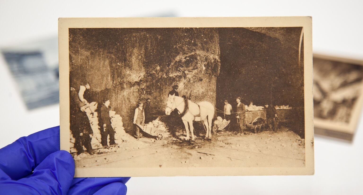 Na dłoni widoczna stare zdjęcie, przedstawiające konia ciągnącego wagoniki. Wokół widać 7 ludzi wykonujących pracę w kopalni.
