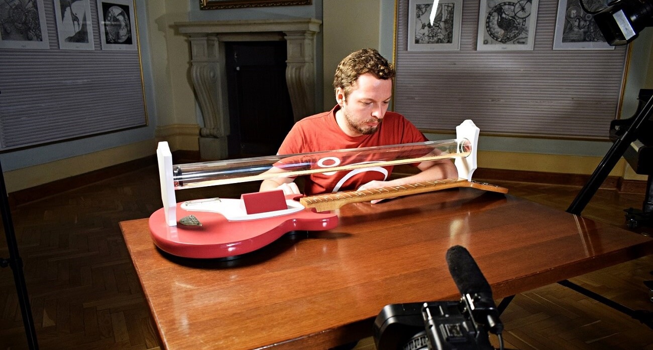 Zdjęcie przedstawia edukatora spoglądającego na gitarę przerobioną na eksponat naukowy. Przed nim widoczny jest fragment kamery, która rejestruje przebieg doświadczenia.