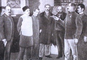 Czarno-białe zdjęcie przedstawiające 7 mężczyzn z których jeden ubrany jest w kamizelkę kuloodporną. Obok niego znajduje się meżczyzna, który celuje do kamizelki z małego pistoletu.